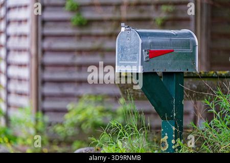 Ein gewölbter Briefkasten aus Metall sitzt auf einem grünen Holzpfosten mit einer roten Flagge. Das kleine Schlüsselloch wartet neben dem Griff, und die ordentliche Box steht bereit für den täglichen Gebrauch Stockfoto