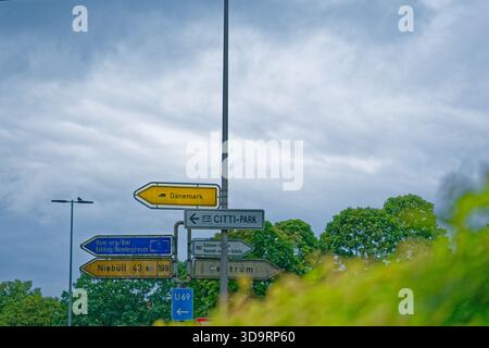 Richtungsschilder bündeln sich an einem Metallmast, links in Richtung Dänemark, Sitti Park in Flensburg, Niebull und Hamburg, rechts in Richtung Zentrum. Bleche in Gelb Stockfoto