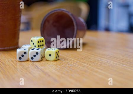Auf der Tischplatte erscheinen zwei Lederbecher, eine aufrecht und eine seitlich. Vier weiße Würfel und zwei blassgelbe Würfel sind verstreut. Sanftes Licht c Stockfoto