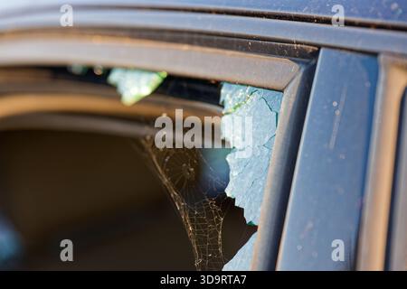 Eine Autotür mit gebrochener Seitenscheibe, feine Teile haften am Rahmen und streuen sich über den Fußraum, Reflexionen glitzern auf dem Glasstaub, das Fahrzeug Stockfoto