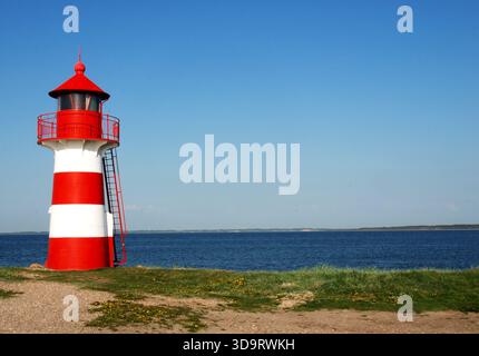 Ein rot-weiß gestreifter Leuchtturm steht an einem grasbewachsenen Ufer am Meer unter einem klaren blauen Himmel. Stockfoto