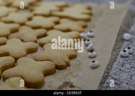 Eine Nahaufnahme von frisch gebackenen Lebkuchenkeksen in Form von Lebkuchenmännern, auf Pergamentpapier gelegt. In der Nähe gibt es Süßigkeitenaugen, die verstreut sind Stockfoto