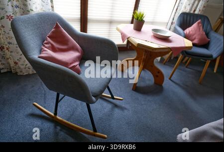 Eine gemütliche Inneneinrichtung mit Schaukelstuhl mit rosa Kissen, Holztisch mit Schüssel und Topfpflanze. Das Zimmer verfügt über sanfte Beleuchtung und fl Stockfoto