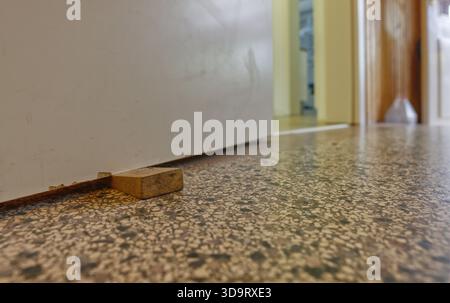 Eine Nahansicht eines hölzernen Türstopps auf einem strukturierten Boden, mit einer Tür, die im Hintergrund sichtbar ist. Die Tür ist leicht offen und offenbart einen Schimmer Stockfoto