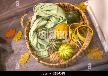 Ein gewebter Korb, gefüllt mit verschiedenen dekorativen Kürbissen und einem hellgrünen Stoff. Um den Korb herum liegen verstreute Herbstblätter, die ein gemütliches fal schaffen Stockfoto