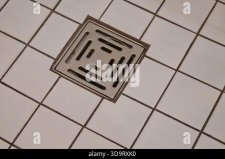Nahansicht einer quadratischen Metallabflussabdeckung auf einem gefliesten Boden mit weißen und grauen Fliesen. Der Abfluss weist ein geometrisches Muster auf und ist umgeben Stockfoto