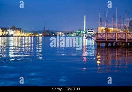 Eine ruhige Küstenszene in der Abenddämmerung mit ruhigem Wasser, das die Lichter der Stadt und die Boote reflektiert. Die Skyline umfasst eine Mischung aus historischem und modernem Architekten Stockfoto
