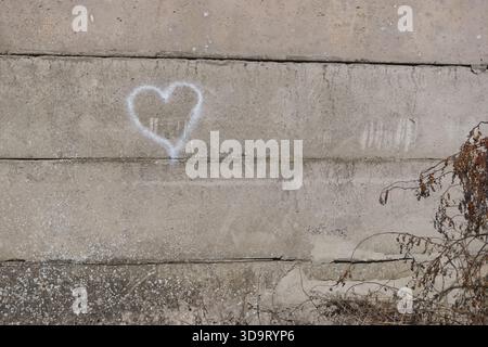 Eine einfache weiße Herzform, die auf eine raue Betonwand gesprüht wurde. Die Wand ist grau und verwittert, mit einigen Flecken weißer Rückstände. Im Vorfeld Stockfoto