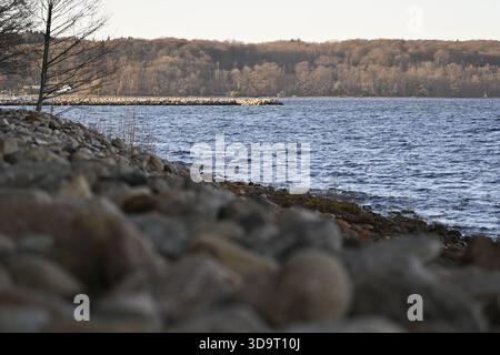 Ein ruhiger Blick auf den See mit einer felsigen Küste, ruhigem Wasser und einem weit entfernten Waldgebiet. Das Sonnenlicht wirft ein warmes Licht auf die Landschaft, Kreatin Stockfoto