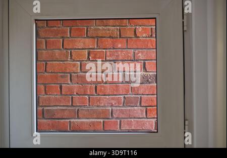 Eine Nahaufnahme einer alten roten Ziegelmauer, die durch einen quadratischen Fensterrahmen gesehen wird. Die Ziegelsteine sind rot mit unterschiedlichen Texturen und Schattierungen, was einen rustikalen ap schafft Stockfoto