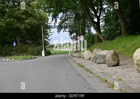 Eine ruhige Straße, die in einen grünen Park führt, gesäumt von großen Steinen auf einer Seite. Bäume bieten Schatten, und im Hintergrund ist ein Wegweiser zu sehen. Th Stockfoto
