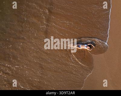 Topdown-Ansicht der jungen Frau, die an der Küste liegt, während Wellen über sie am Talalla Beach, Sri Lanka, strömen Stockfoto