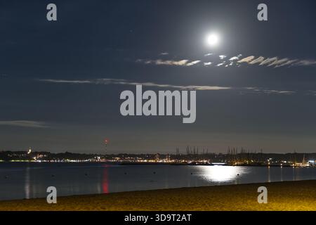 Eine ruhige Nachtszene mit einem ruhigen Strand mit mondbeleuchtetem Himmel. Der Mond reflektiert sich auf dem Wasser und beleuchtet die Küste. Im Hintergrund, City li Stockfoto