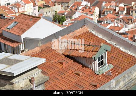 Blick auf ein Dach mit Terrakottafliesen und ein kleines Fenster, das aus dem schrägen Dach ragt. Eine Scheibe ist kaputt und das Glas liegt auf dem Dach i Stockfoto