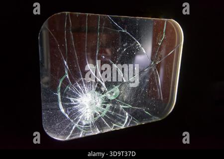 Nahaufnahme einer Glasscheibe mit spinnennetzähnlichem Rissmuster und zentraler Aufprallstelle. Das Glas wird vor einem dunklen Hintergrund beleuchtet Stockfoto