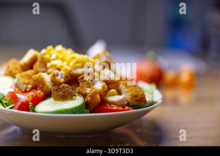 Eine Nahaufnahme eines bunten Salats mit knusprigen Hühnchenstückchen, geriebenem Käse, geschnittenen Gurken und Kirschtomaten, serviert in einer weißen Schüssel auf einem wo Stockfoto