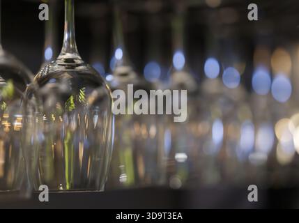 Eine Reihe von Weingläsern, die kopfüber hängen und ihre eleganten Formen und Reflexionen zeigen. Der Hintergrund ist mit weichem Bokeh-Licht versehen Stockfoto