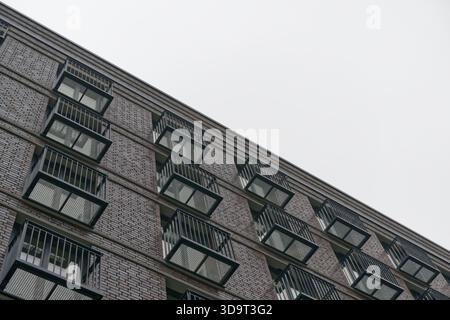 Ein flacher Blick auf ein modernes Backsteingebäude mit mehreren Balkonen mit Metallgeländern. Der Himmel ist bedeckt und schafft eine stimmungsvolle Atmosphäre. Stockfoto