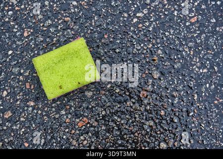 Ein grüner Schwamm, der auf einer nassen, strukturierten Oberfläche mit kleinen Steinen und Kieselsteinen liegt. Der Schwamm hat einen violetten Rand und ist von einem glänzenden, reflektierenden s umgeben Stockfoto