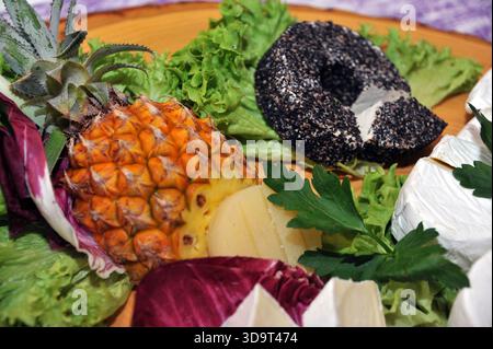 Eine lebendige Platte mit frischem Obst und Gemüse, einschließlich Ananas, Blattgemüse und einem Schwarzkäserad, die künstlerisch auf einem Wald angeordnet ist Stockfoto