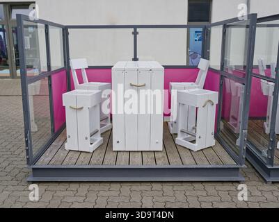 Ein moderner Sitzbereich im Freien mit weißen Holzmöbeln, einschließlich zwei Stühlen und einem Tisch, umgeben von Glaspaneelen mit rosa Akzenten. Th Stockfoto