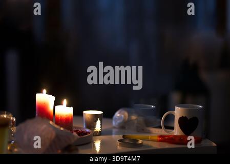 Eine gemütliche, schwach beleuchtete Szene mit drei Kerzen, eine mit Weihnachtsbaummotiv und einem weißen Becher mit Herz. Der Tisch ist mit ein paar Dekorationen geschmückt Stockfoto