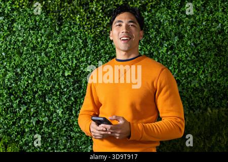 Asiatischer Mann in 20ern, der vor Efeuhecke ein orangefarbenes Sweatshirt trägt, das Smartphone hält und nach oben blickt Stockfoto