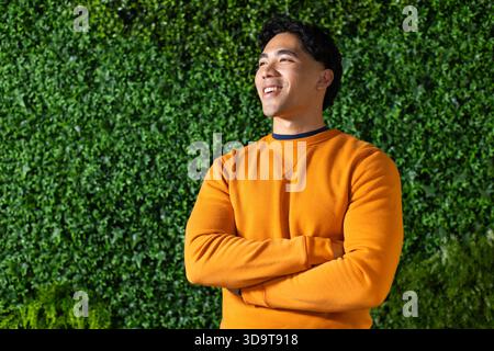 Asiatischer Mann, der lächelnd mit überkreuzten Armen in orangefarbenem Sweatshirt an der Heckenwand steht, Kopierraum Stockfoto