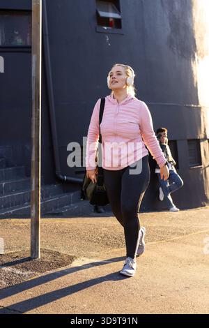Frau, die auf dem Gehweg neben einem grauen Gebäude läuft, trägt einen rosafarbenen Hoodie mit Turntasche und Kopfhörern Stockfoto