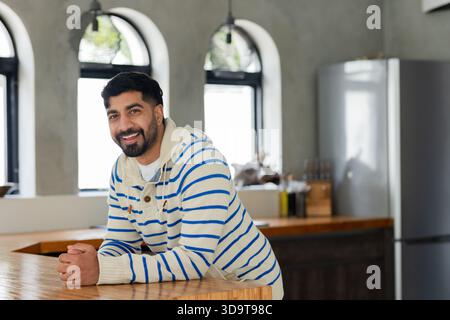 Indischer Mann lehnte sich auf eine hölzerne Küchenarbeitsfläche und lächelte neben Olivenölflaschen und Kühlschrank Stockfoto