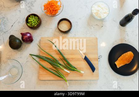 Holzbrett mit grünen Zwiebeln und blauem Messer auf Marmorarbeitsfläche mit Schüsseln und Lachs Stockfoto