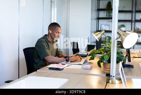 Afroamerikanischer Mann, der am Schreibtisch sitzt und auf dem Laptop schreibt, Ohrstöpsel trägt und ein Notebook benutzt Stockfoto