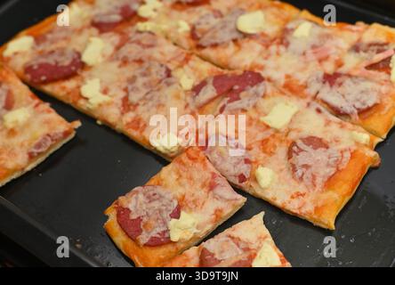 Heiße Pizza mit Peperoni und Käse, frisch gebacken auf einem schwarzen Backblech. Die Pizza wird in rechteckige Scheiben geschnitten und kann serviert werden. Stockfoto