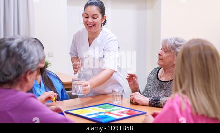 Pflegekraft, die Seniorinnen beim Brettspiel Wasser gibt Stockfoto