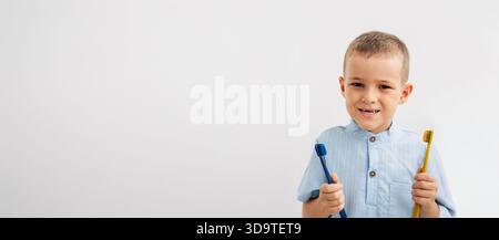 Lächelnder Junge mit einer natürlichen Zahnlücke, der zwei bunte Zahnbürsten auf weißem Hintergrund hält Stockfoto