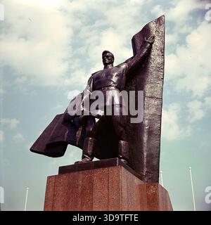 Ein Vintage-Farbfoto im Archivformat, das das riesige Kriegsdenkmal in Slowiansk, Donezk, Ukraine, während der 1980er Jahre festgehalten hat Die dunkle Metallskulptur zeigt einen sowjetischen Soldaten, der ein Maschinengewehr und ein Banner vor einem blauen Himmel hält. Diese dramatische, aus einem tiefen Winkel gezogene Aufnahme im Lenin Park (heute Silkovichny Park) unterstreicht den heldenhaften Stil des sozialistischen Realismus. Das Denkmal steht als mächtiges Symbol der Erinnerung an den Zweiten Weltkrieg und der Geschichte in der sonnigen Atmosphäre eines friedlichen Donbas. Stockfoto