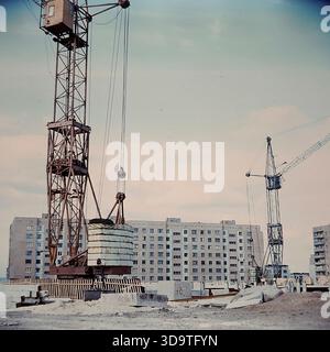 Vintage-Archivfoto aus den 1980er Jahren mit einer großen Baustelle in Sloviansk, UdSSR. Turmkräne erheben sich über einem neuen Wohnviertel und bauen ein mehrstöckiges Paneelgebäude aus Betonplatten zusammen. Der Vordergrund ist mit Baumaterialien und Fundamentblöcken gefüllt, die auf dem Sandboden verstreut sind. Diese Industrielandschaft veranschaulicht die rasante städtische Entwicklung, die Erweiterung des Wohnraums und die architektonische Transformation der friedlichen Donbass-Region während der späten Sowjetzeit. Stockfoto