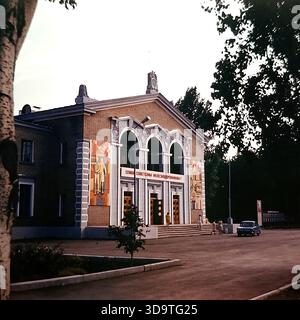 Vintage-Archivfoto aus den 1980er Jahren, in dem der Kulturpalast der Eisenbahnarbeiter in Sloviansk, UdSSR, aufgenommen wurde. Das Backsteingebäude verfügt über drei große Bögen und farbenfrohe Mosaikpaneele an der Fassade. Ein Slogan: „Ruhm an die sowjetischen Eisenbahnarbeiter!“ Hängt über dem Eingang. Ein blaues sowjetisches Auto parkt in der Nähe, und Einheimische gehen über den Platz. Eingerahmt von Bäumen spiegelt dieses authentische Bild das kulturelle Leben und die Bedeutung der Eisenbahnindustrie im friedlichen Donbass wider. Stockfoto