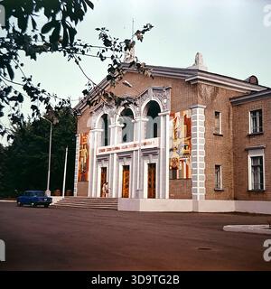 Vintage-Archivfoto aus den 1980er Jahren, in dem der Kulturpalast der Eisenbahnarbeiter in Sloviansk, UdSSR, aufgenommen wurde. Das Backsteingebäude verfügt über drei große Bögen und farbenfrohe Mosaikpaneele an der Fassade. Ein Slogan: „Ruhm an die sowjetischen Eisenbahnarbeiter!“ Hängt über dem Eingang. Ein blaues sowjetisches Auto parkt in der Nähe, und Einheimische gehen über den Platz. Eingerahmt von Bäumen spiegelt dieses authentische Bild das kulturelle Leben und die Bedeutung der Eisenbahnindustrie im friedlichen Donbass wider. Stockfoto