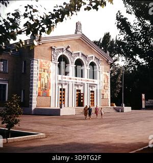 Vintage-Archivfoto aus den 1980er Jahren, in dem der Kulturpalast der Eisenbahnarbeiter in Sloviansk, UdSSR, aufgenommen wurde. Das Backsteingebäude verfügt über drei große Bögen und farbenfrohe Mosaikpaneele an der Fassade. Ein Slogan: „Ruhm an die sowjetischen Eisenbahnarbeiter!“ Hängt über dem Eingang. Ein blaues sowjetisches Auto parkt in der Nähe, und Einheimische gehen über den Platz. Eingerahmt von Bäumen spiegelt dieses authentische Bild das kulturelle Leben und die Bedeutung der Eisenbahnindustrie im friedlichen Donbass wider. Stockfoto