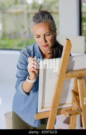 Frau mittleren Alters malt auf Leinwand an der Staffelei im Studio und hält Pinsel in der Nähe der Smartwatch Stockfoto