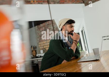 Indischer Mann lehnt sich auf eine hölzerne Küchentheke und starrt auf silbernen Laptop mit Keramikbecher am Fenster Stockfoto