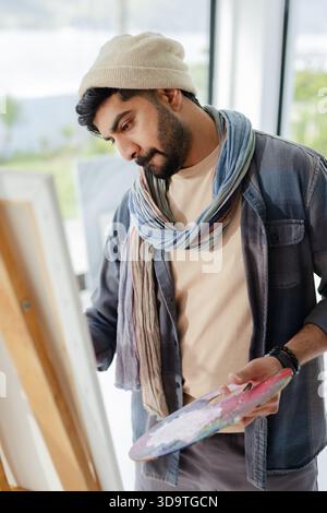 Indischer Mann malte auf Leinwand an der Staffelei im Kunststudio, hielt Palette, Pinsel, trug Strickmütze Stockfoto