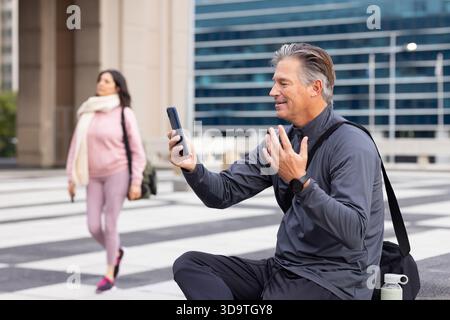 Reifer Mann, der am plaza Ledge ein Smartphone anruft, reife Frau, die mit Tasche in der Nähe eines Glasgebäudes läuft Stockfoto