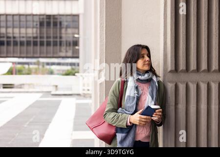 Frau, die sich gegen eine Betonsäule auf dem städtischen platz lehnt und Kaffeetasse und Smartphone hält, Kopierraum Stockfoto