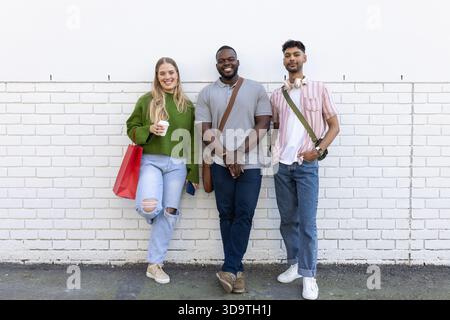 Verschiedene Freunde, die sich an der Wand lehnen und eine Kaffeetasse, eine rote Tasche, ein Smartphone, eine Messenger-Tasche und Kopfhörer halten Stockfoto