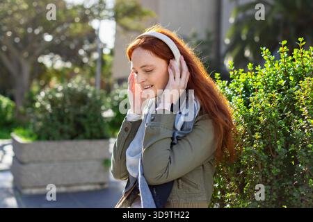 Frau, die schnurlose Kopfhörer auf dem Gehweg neben dem Pflanzgefäß anpasst, trägt grüne Jacke und Smartwatch Stockfoto