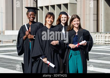 Verschiedene weibliche Absolventen stehen auf dem gefliesten Campus plaza in Gewändern und halten Diplome mit roten Bändern Stockfoto