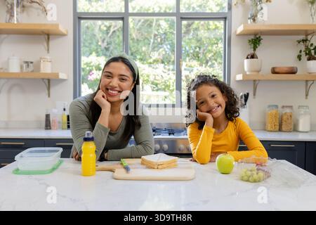 Mutter und Tochter lächeln, während sie sich auf die Arbeitsplatte in der Küche mit hölzernem Schneidebrett lehnen Stockfoto