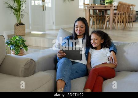 Mutter und Tochter sitzen auf hellgrauem Sofa im Wohnzimmer und teilen sich den Tablet-Computer neben der Pflanze Stockfoto